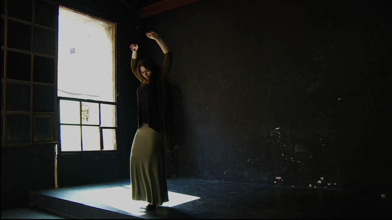 A young women in a dim studio practices flamenco in a long taupe skirt and dark sweater